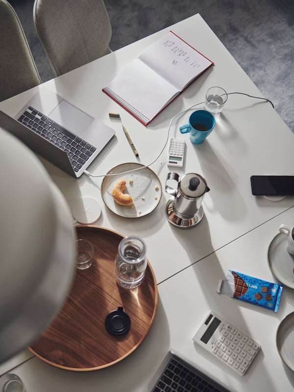 A table formed by two side-by-side white TROTTEN desks, scattered with office and coffee-break accessories.
