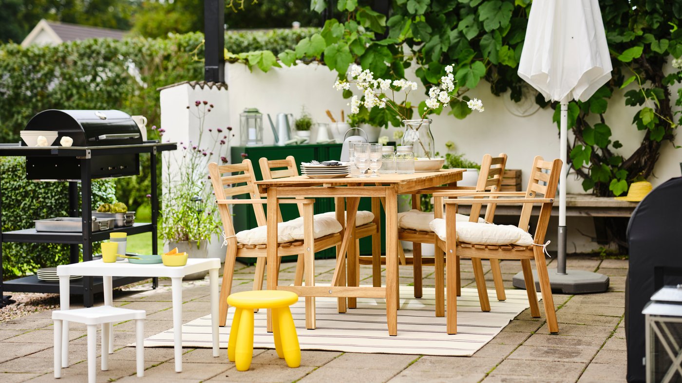IKEA Terrassenszene mit NÄMMARÖ Gartentisch, vier Holzstühlen mit weißen Polstern, Kinder-Sitzgruppe und Grill, umgeben von Pflanzen und Sommerdeko.