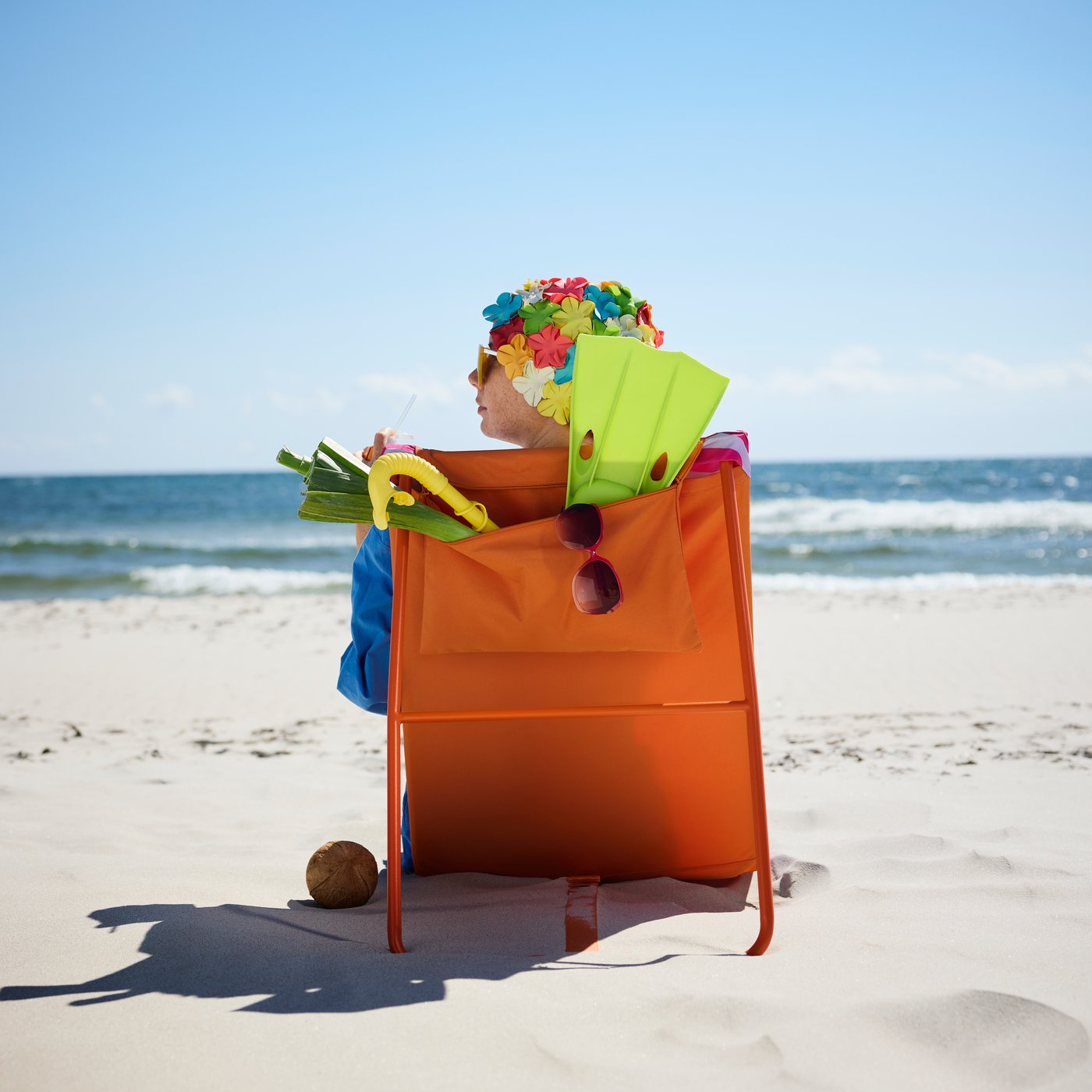 Eine Dünenlandschaft mit einer bunten STRANDÖN Sonnenliege, auf der eine Person mit einer geblümten Schwimmhaube in die Ferne schaut