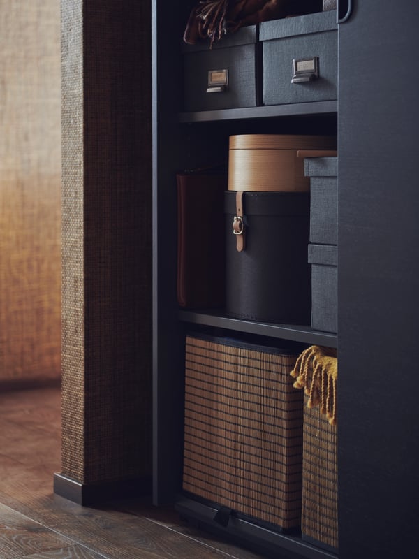 A black GALANT cabinet with one sliding door open and three shelves holding storage boxes with lids, round boxes and baskets.