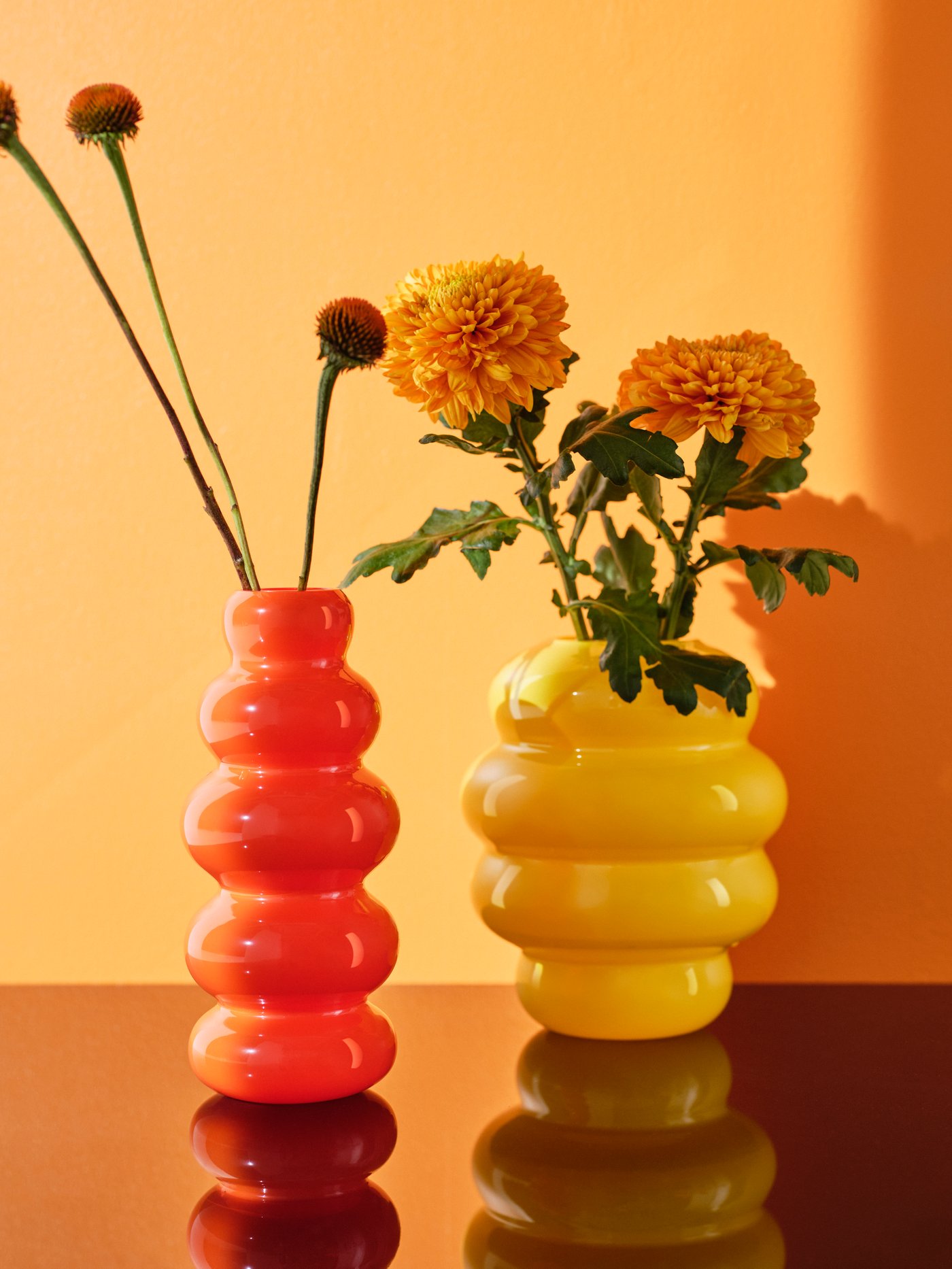Fleurs dans deux vases KÄLLARHALS, un orange et un jaune, posés sur une surface brillante dans laquelle ils se reflètent.