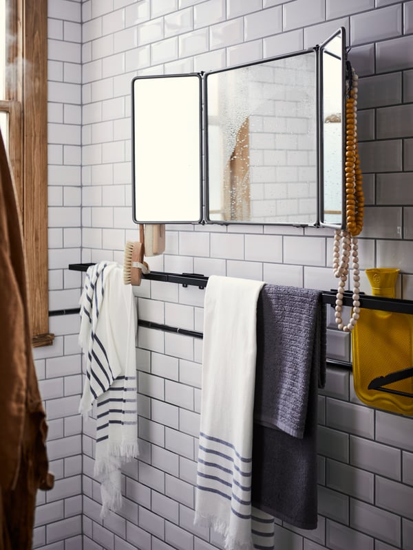 SKOGSVIKEN black towel rail with towels against a white tile bathroom wall with a SYNNERBY tri-fold mirror above.