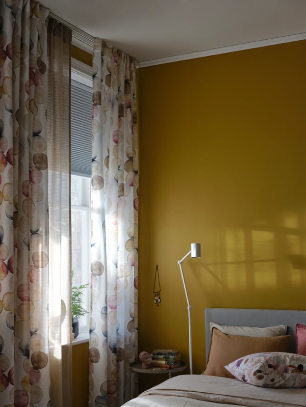 Krevet i podna/čitaća lampa pored prozora sa zavesama na belim VIDGA 1-kanalnim garnišnama i neprozirna ćelijasta roletna.