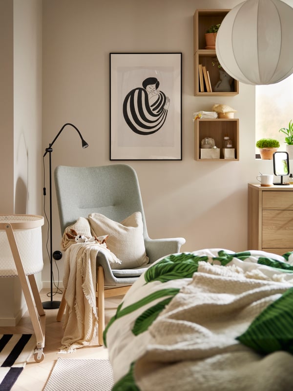 VEDBO fauteuil met hoge rugleuning en een plaid over een van de armleuningen staat in een hoek naast een bed en een ÄLSKVÄRD wieg met matras.