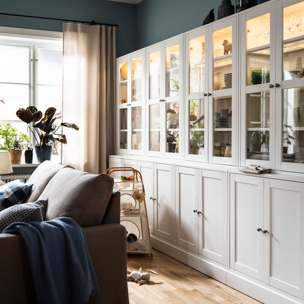 Some white HAVSTA storage combinations with glass-doors stand against a wall behind a grey-brown HYLTARP sofa-bed.