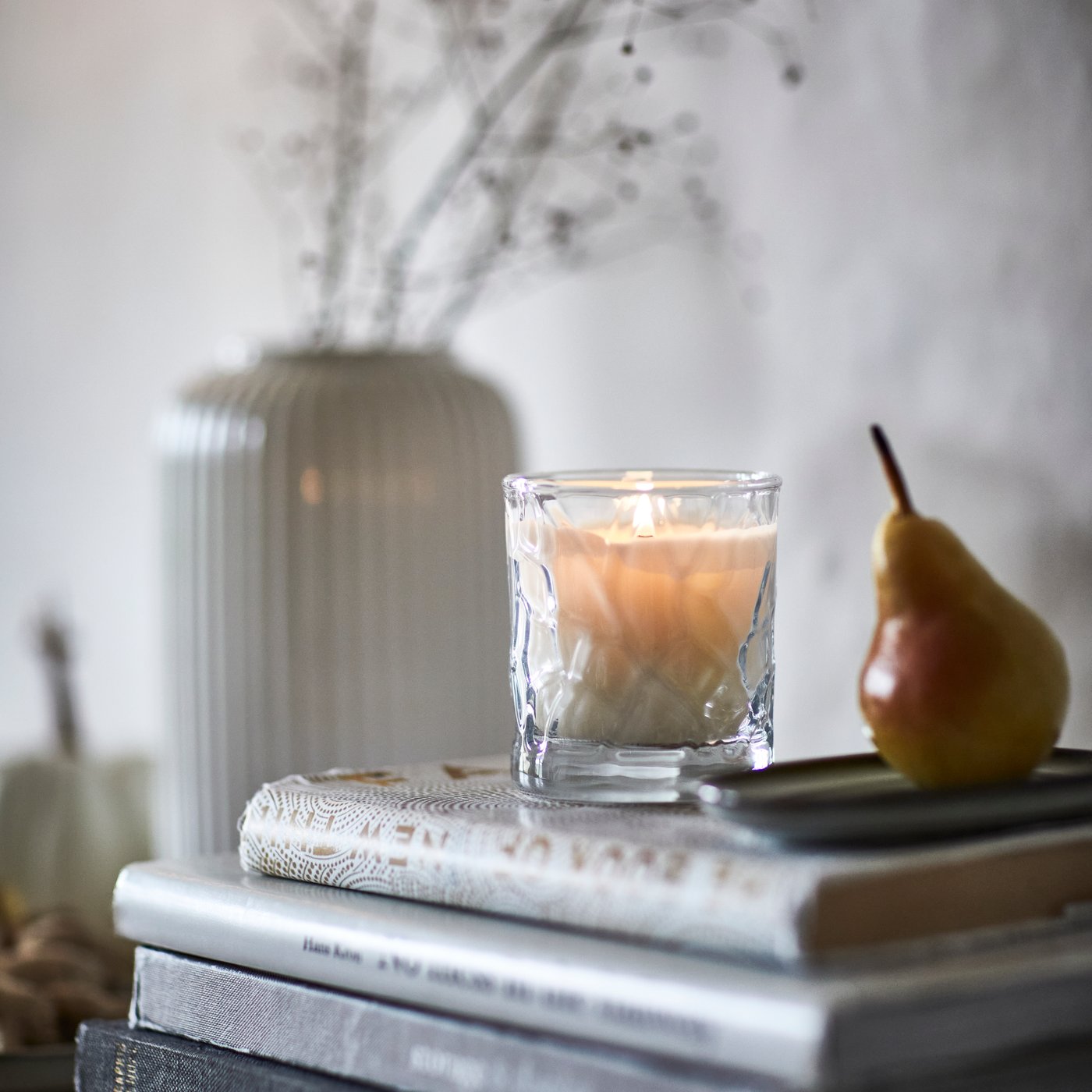 Una candela profumata HÖSTAGILLE in un barattolo di vetro è appoggiata su una pila di libri accanto a un vaso decorativo e una pera.