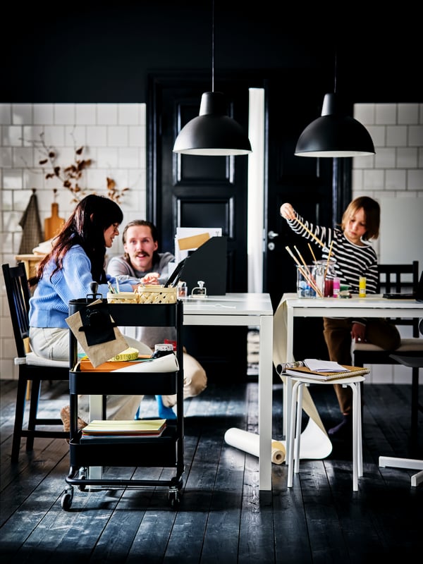Family members doing parallel activities by white MELLTORP tables and a black RÅSKOG cart in a black-and-white kitchen.