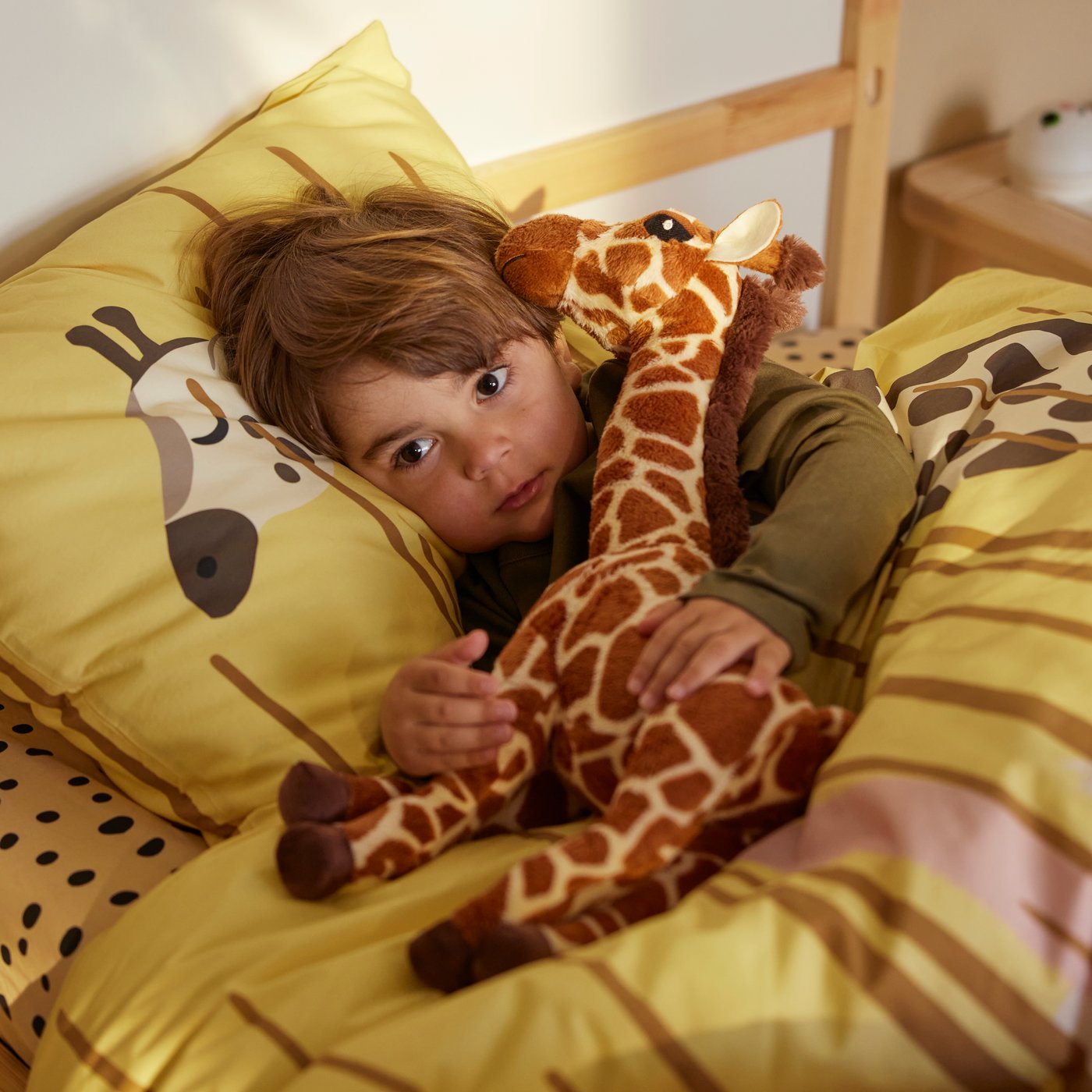 A child is cuddling with a SANDLÖPARE soft toy giraffe calf in a bed with yellow SANDLÖPARE duvet cover and pillowcase.