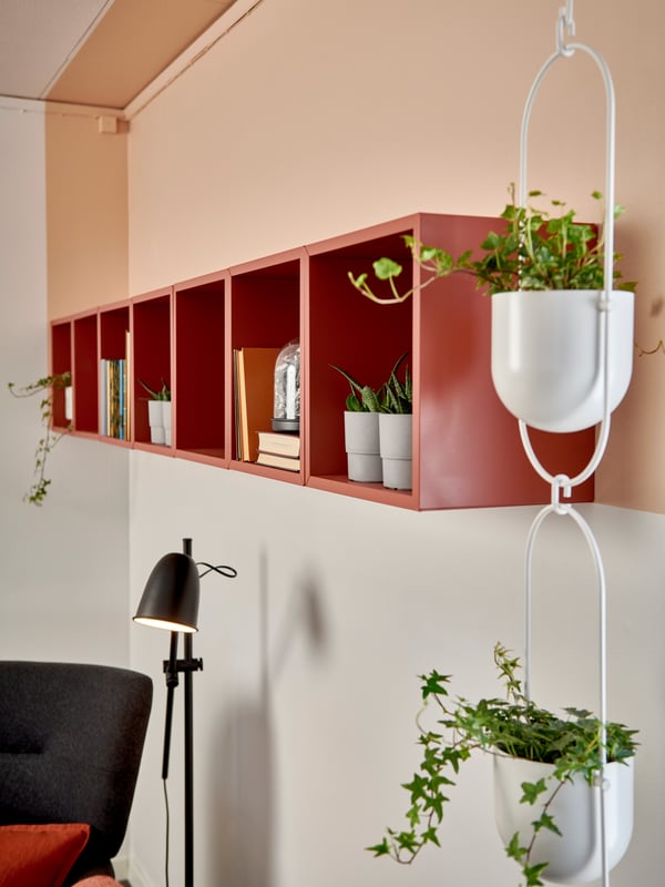 Röd hylla med böcker och växter på ljus vägg, svart golvlampa och två vita hängkrukor med gröna växter.