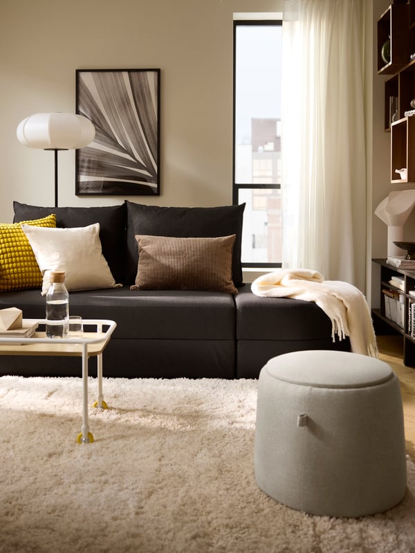 Helles Zimmer mit einem dunkelgrauen LINDÅKRA Bettsofa, einem JÄRRESTAD Pouf, Textilien und einem weissen VOLLERSLEV Teppich.