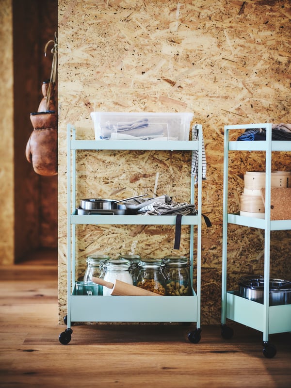 Two pale green NISSAFORS utility carts holding glass storage jars of food and stacked pots and pans sit near a woodchip wall.