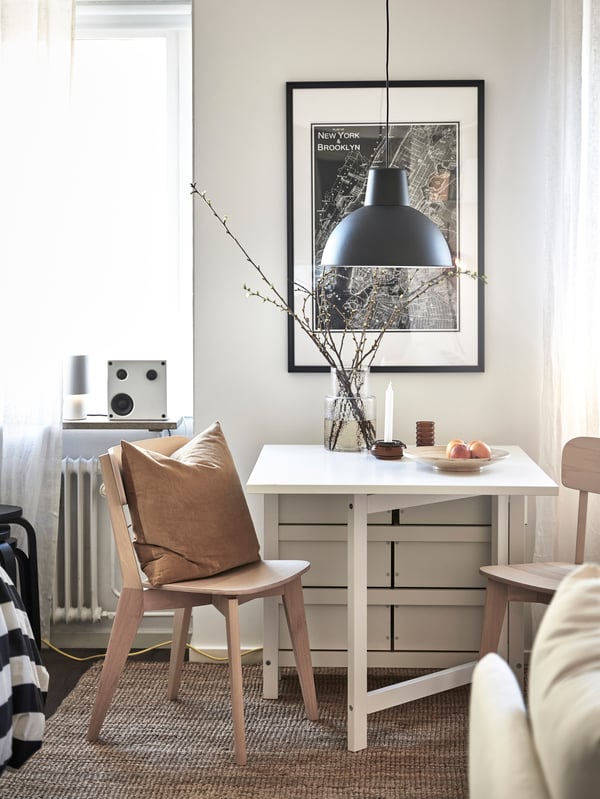 En smart inredd etta med en flexibel matplats med ett vitt slagbord och två stolar i ljust trä. Ovanför matbordet hänger en svart lampa och bakom syns en stor tavla med stadsobjekt.