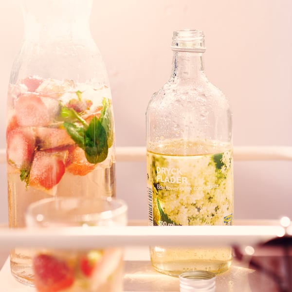 A carafe filled with water, strawberries, and mint leaves stands next to a bottle labeled “DRYCK FLÄDER” filled with a yellow liquid.