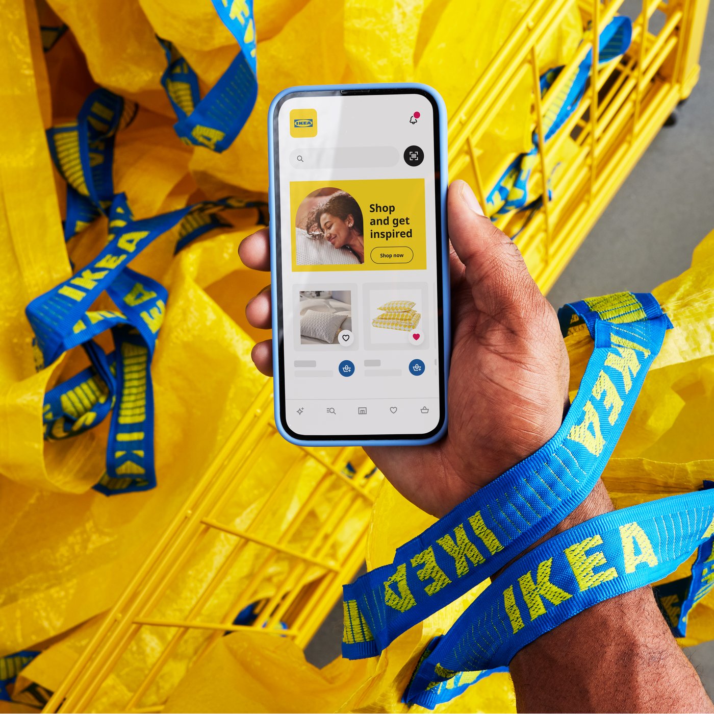 A hand holds a smartphone displaying the IKEA app, surrounded by yellow IKEA bags with blue handles in a shopping cart.