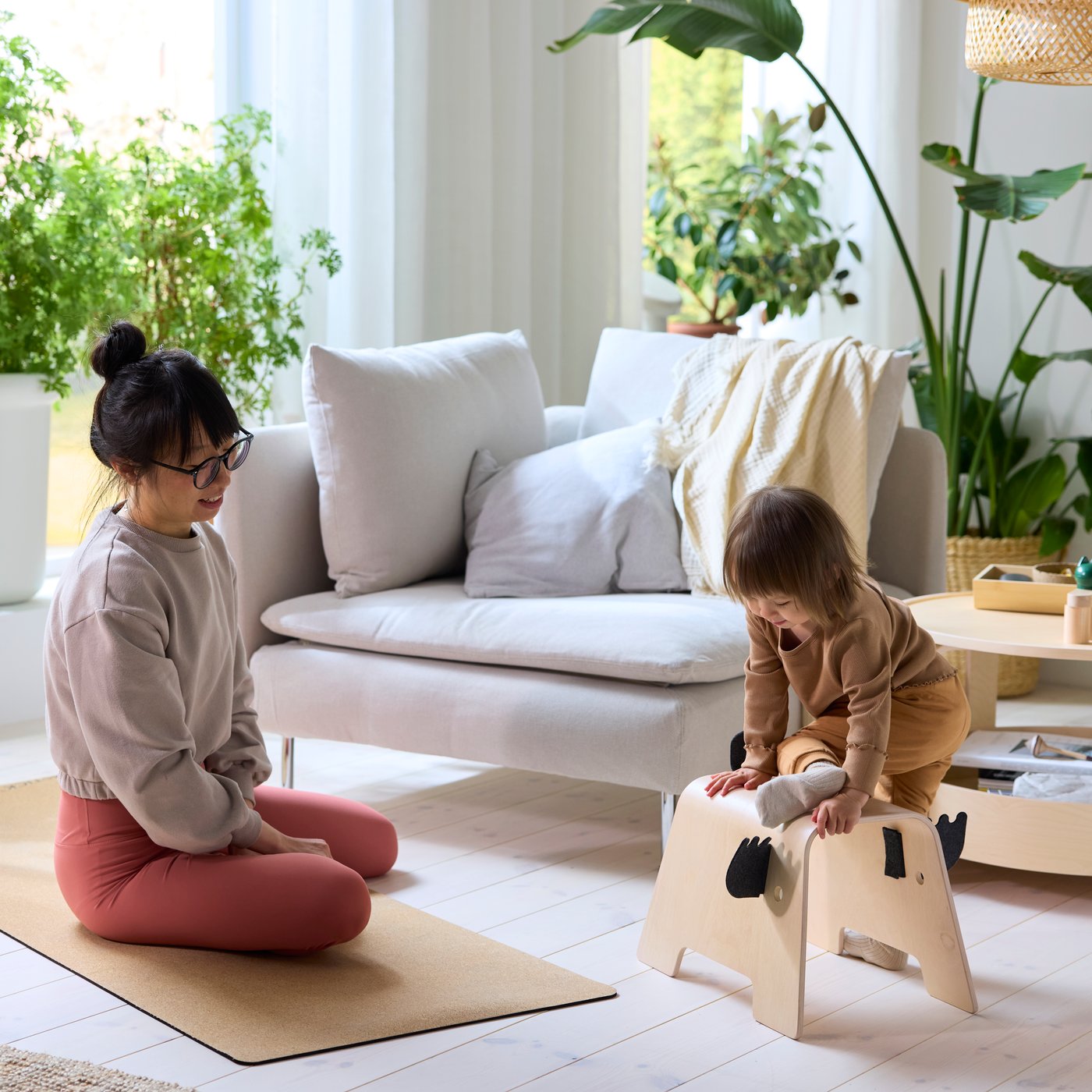 A person sitting on a yoga mat watches their child plays on a GREJSIMOJS children’s stool in a bright living room.