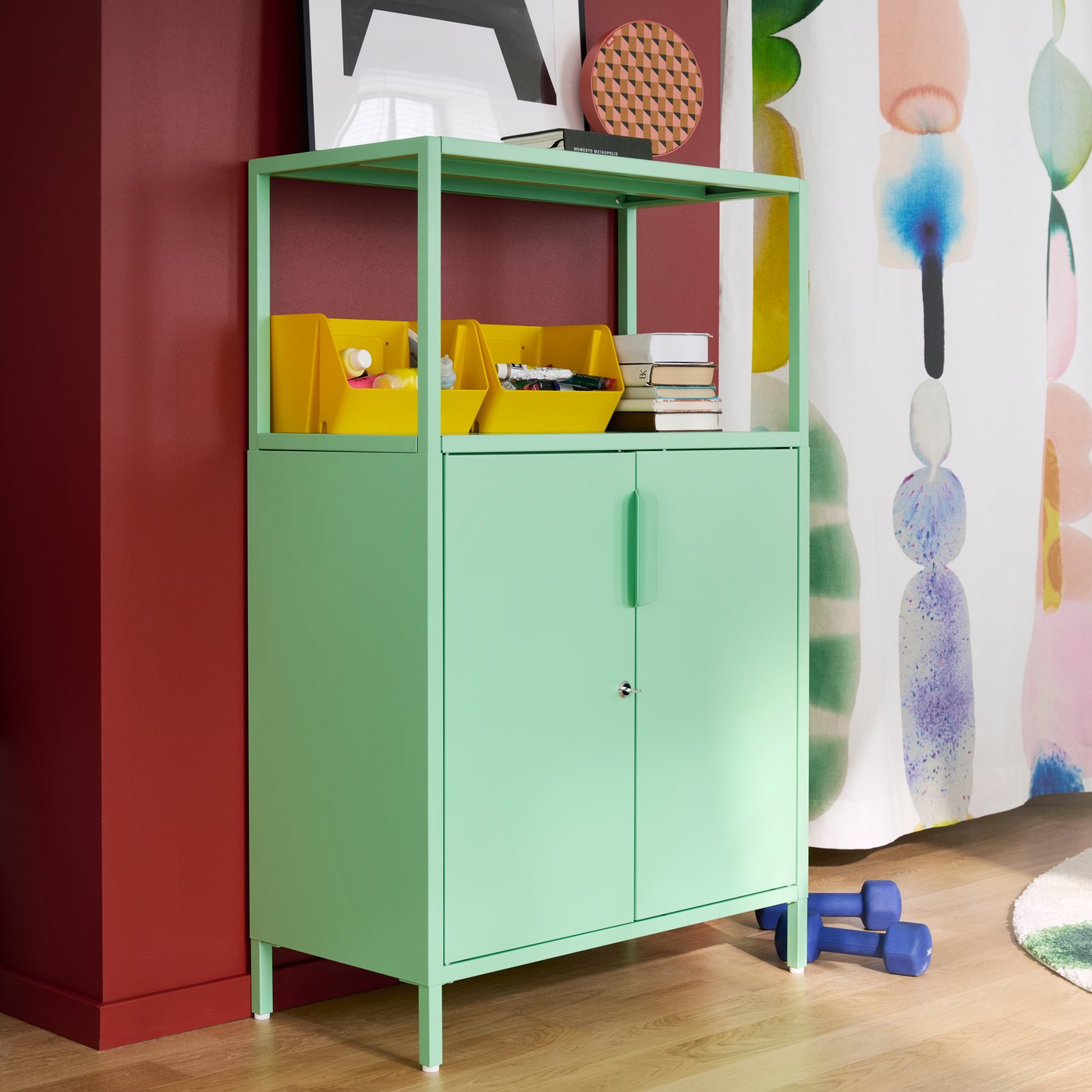 A bright room features a light green TROTTEN cabinet with doors, holding supplies and books beside colourful wall art.