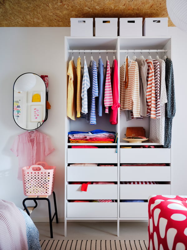 A white AURDAL wardrobe combining closed and open storage, filled with colorful garments. A LINDBYN mirror hangs next to it.