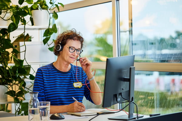Eine Person in IKEA Uniform arbeitet an einem Schreibtisch mit Headset und Notizblock vor einem Computerbildschirm in einem hellen, pflanzenreichen Arbeitsbereich.