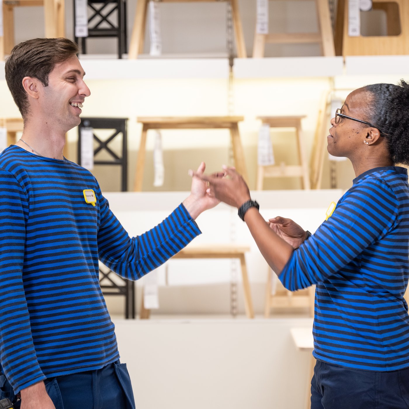 Twee IKEA medewerkers dragen blauw gestreepte shirts en geven elkaar de hand in een IKEA showroom met stoelen op de achtergrond.