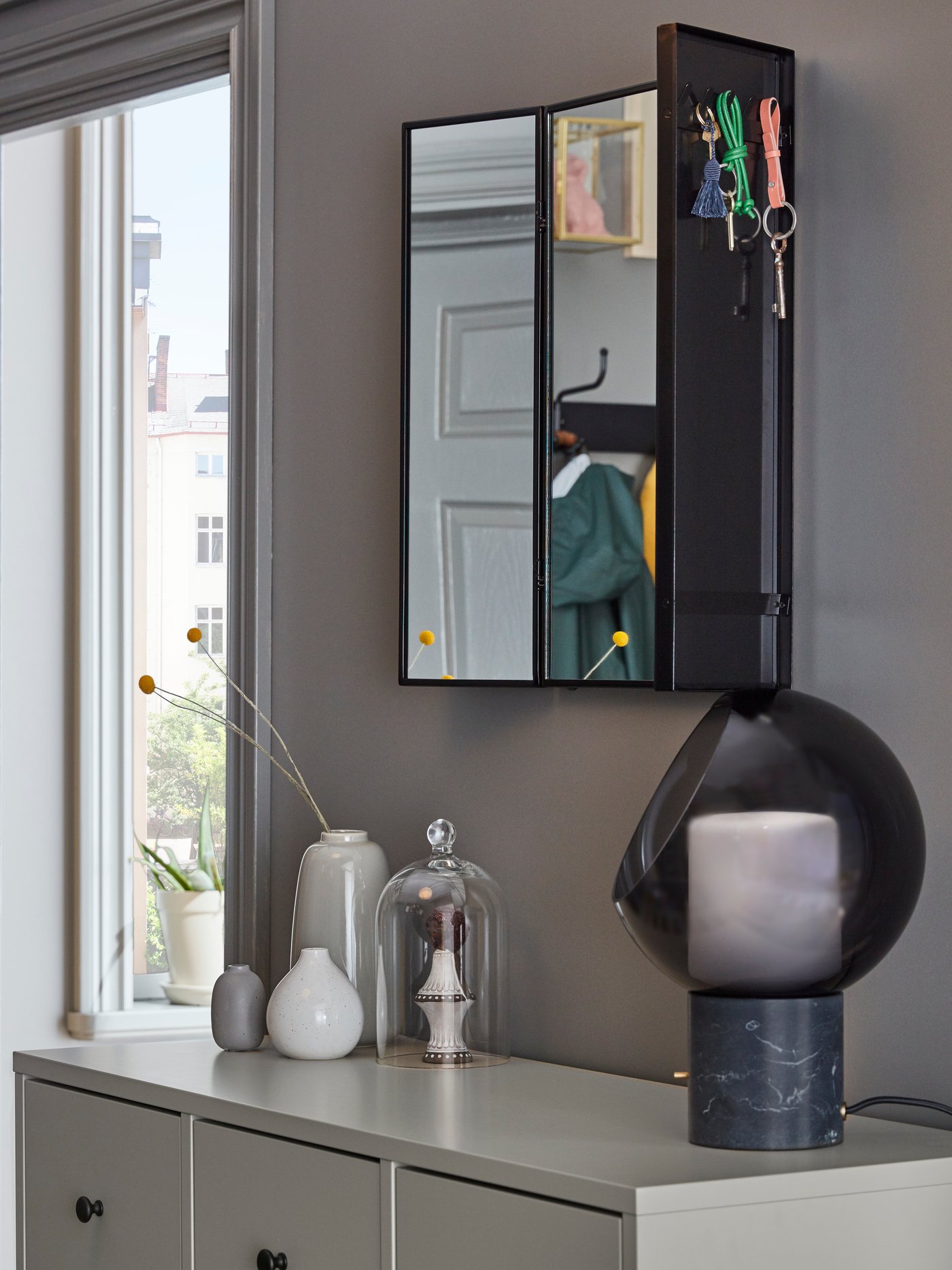 A KARMSUND mirror on a wall with hooks at the back holding keys, above a BRYGGJA chest of drawers with an EVEDAL lamp on it.