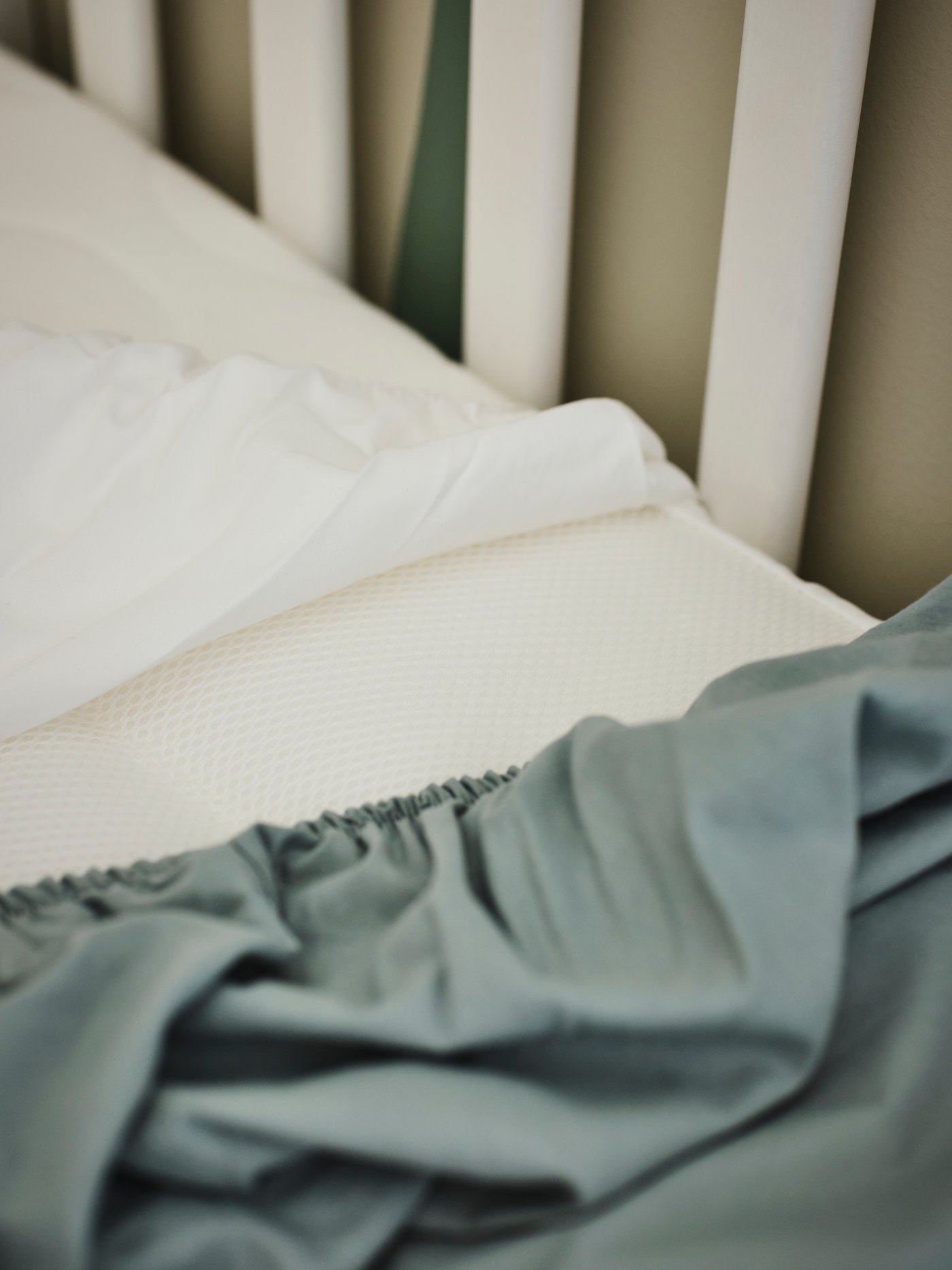 A SKÖNAST foam cot mattress in a white SUNDVIK cot with a ruffled blue GULDVÄVARE fitted sheet on top of it.