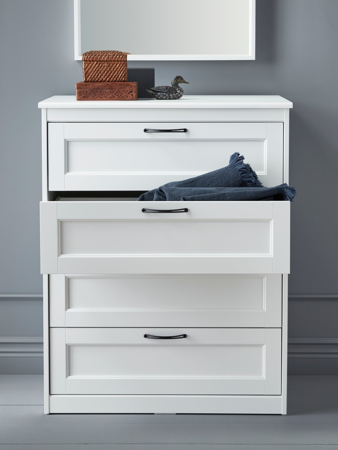 Une commode blanche à trois tiroirs laisse entrevoir un jean plié dans le tiroir du milieu, avec deux paniers en osier et un petit canard décoratif posés dessus.