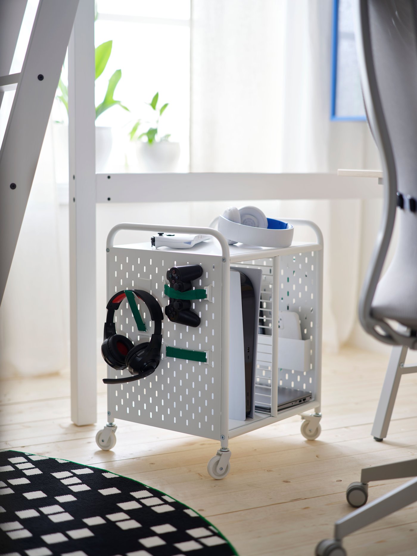 A white BRÄNNBOLL shelving unit on castors holds a headset and controllers on its pegboard side, by a rug and a gaming chair.