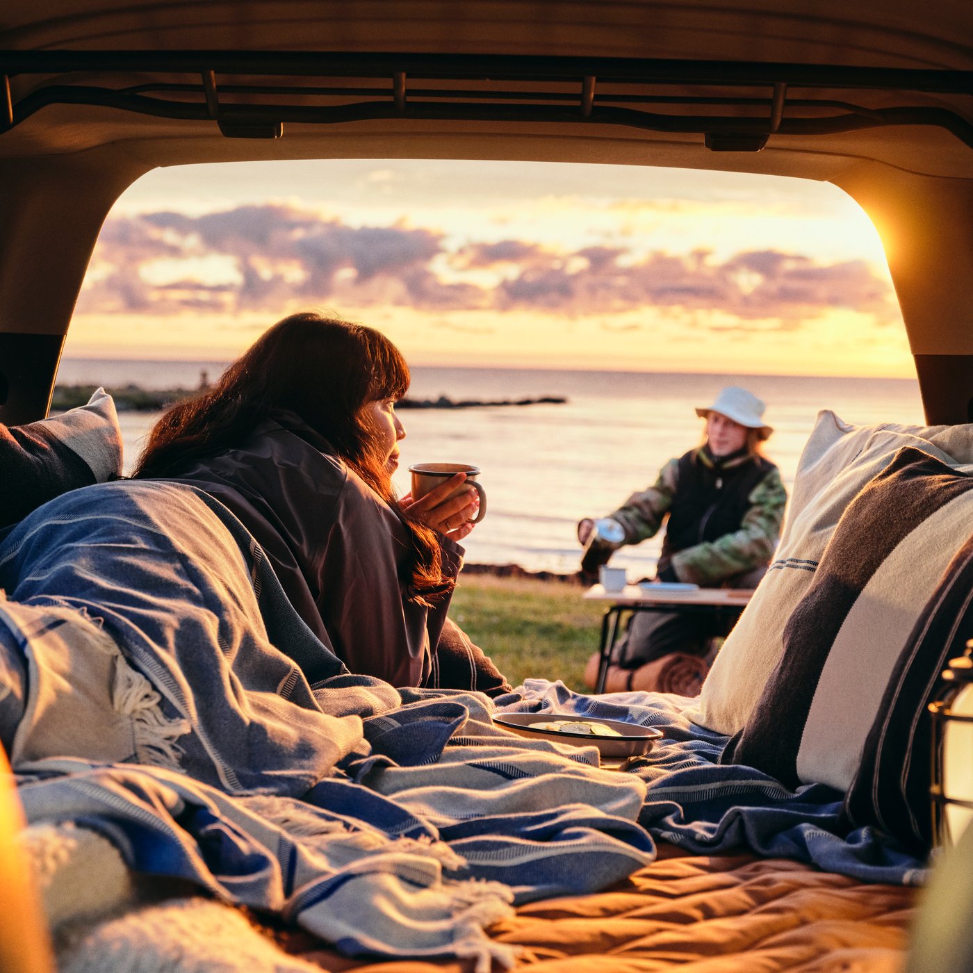 A scenic outdoor setting with a person in a SOLUPPGÅNG throw enjoying a hot drink at the back of a car, overlooking water.
