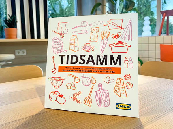 Couverture du livre de recettes TIDSAMM avec illustrations d’ingrédients et d’ustensiles de cuisine sur une table dans un environnement IKEA.