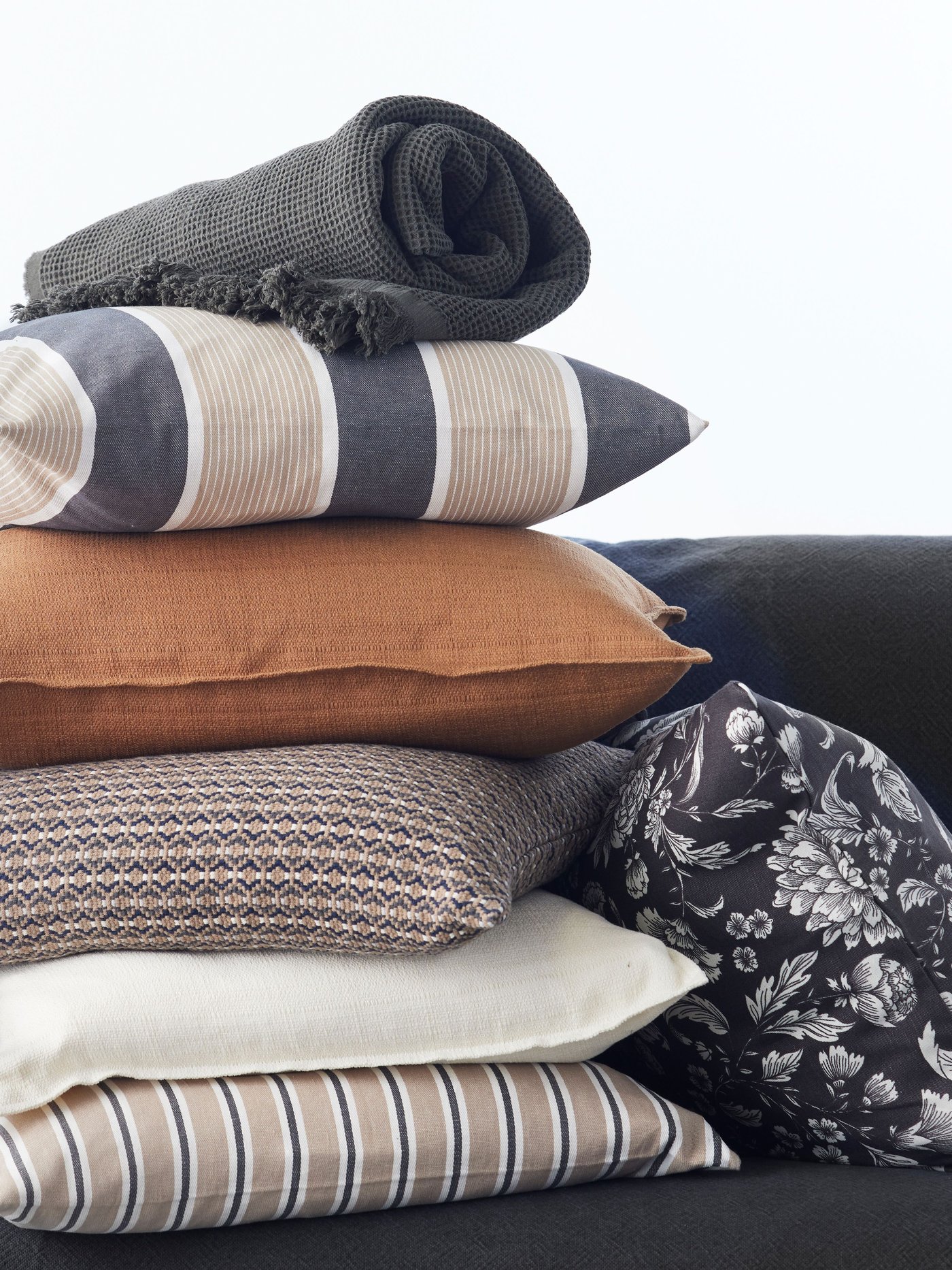 Hromada dekorativních polštářů IKEA v různých vzorech a barvách, na vrchu složená šedá pletená deka.