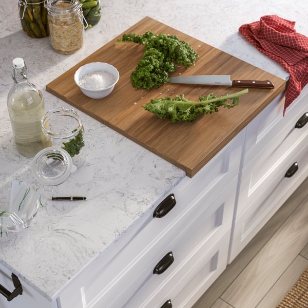 Une planche à découper avec du chou frisé frais et un couteau sur un comptoir de cuisine en quartz, à côté d’une bouteille et de bocaux.