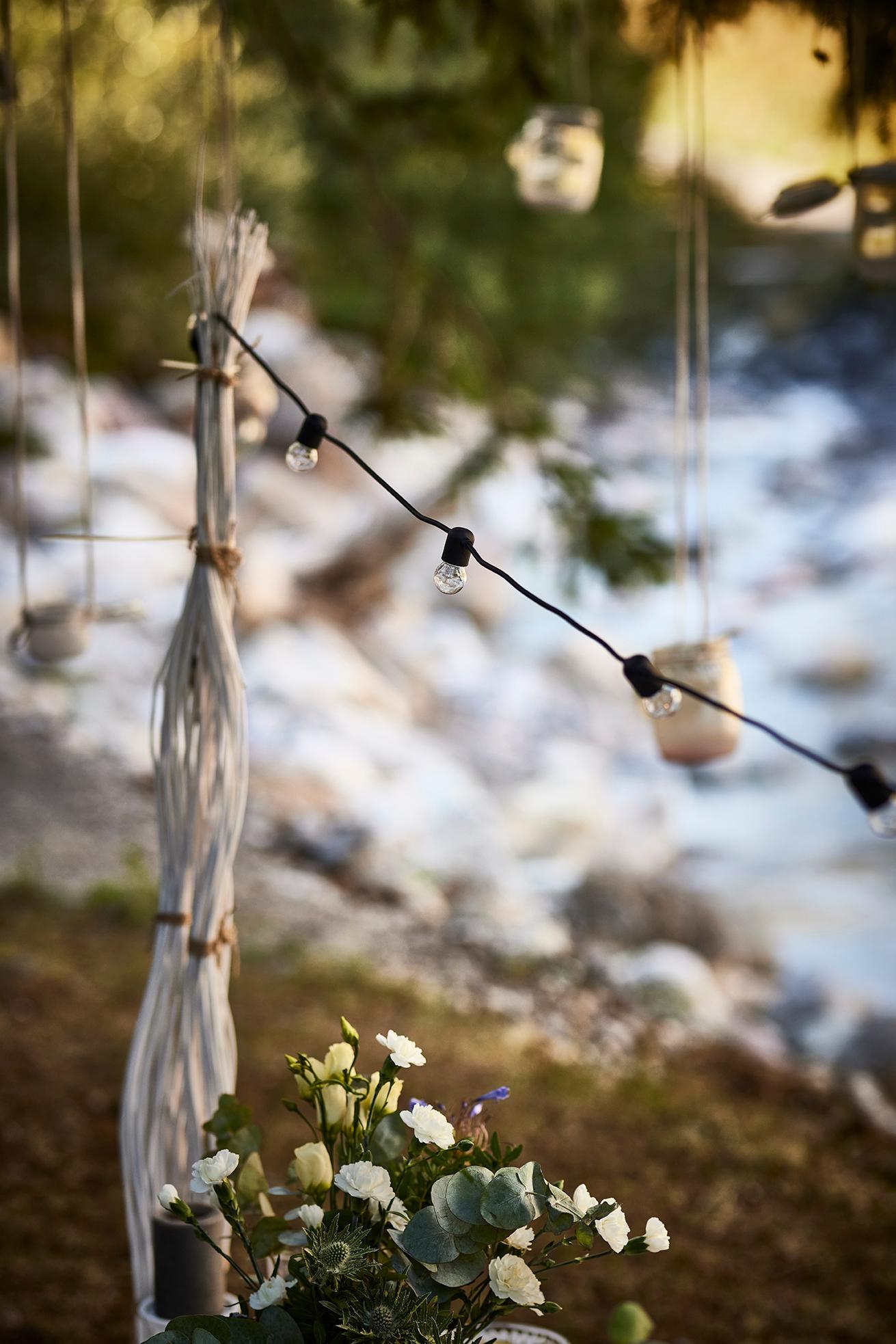 Outdoor-Dekoration mit Blumen, Lichterkette und hängenden Windlichtern am Flussufer für stimmungsvolle Atmosphäre.