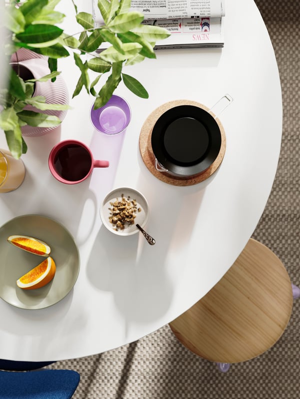 Ett DOCKSTA dukat frukostbord med kaffe, frukt och skålar, bredvid en pall och en krukväxt i bakgrunden.
