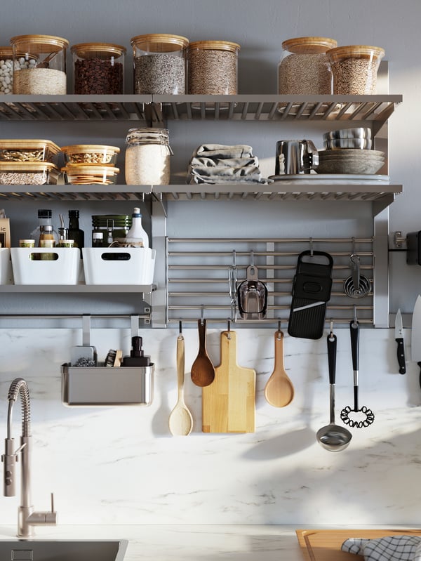 La imagen muestra tres niveles de repisas de cocina metálicas. En los niveles superiores hay frascos transparentes con especias y alimentos no perecibles, mientras que en el nivel inferior se ve un organizador KUNGSFORS con tablas, cucharas y cucharones colgando. 