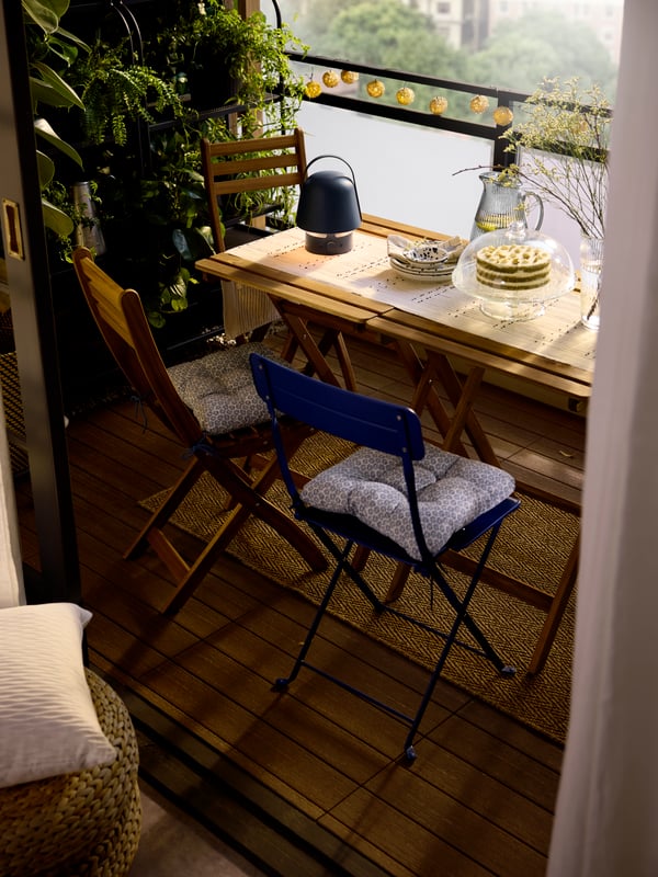 Bujan balkon s malim stolicama i stolovima, na kojima su plavi baštenski VAPPEBY Bluetooth zvučnik-lampa, tanjiri i stalak za tortu.