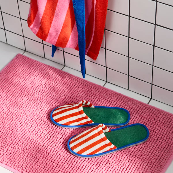 Two SKOGSSALLAT slippers with red and white stripes on the outside, green and blue on the inside. Pink bath mat, white floor.