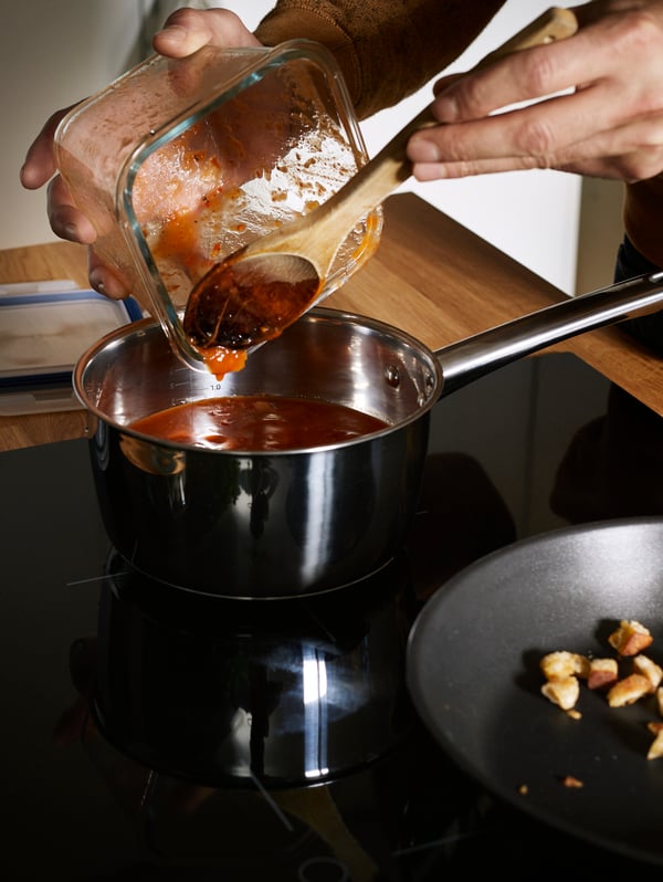 The hands of a person using a wooden spoon to scrape tomato sauce from a square glass food container into a saucepan.