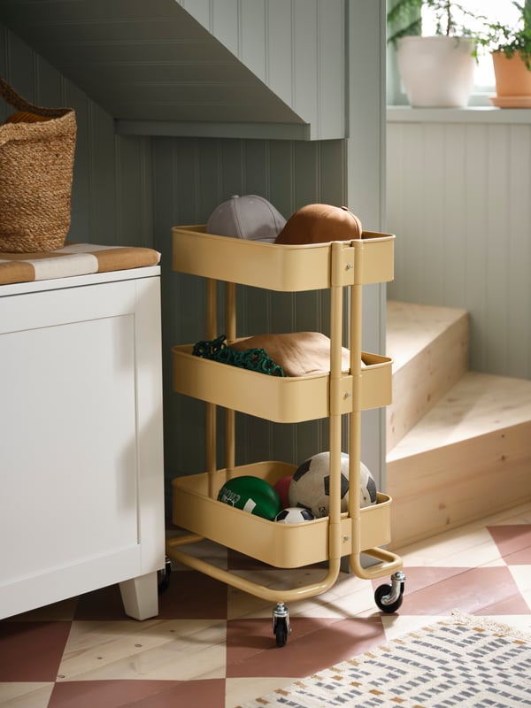 Ein gelber RÅSKOG Servierwagen steht vor einem Teil eines weißen PLATSA Kleiderschranks, der sich unter der Treppe eines Hauses befindet. 