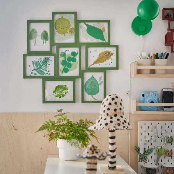A room with a green VÄXBO collage frame for eight drawings arranged on a white wall above plant-filled shelves.