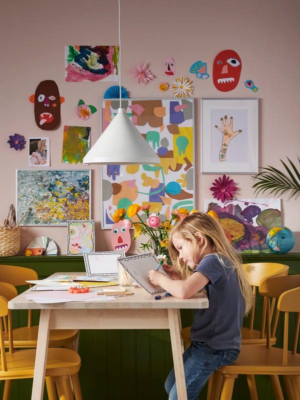 In einem Raum hängen viele bunte Bilder an einer Wand und davor sitzt 1 kleines Kind an einem Tisch und zeichnet etwas auf eine weiße Tafel.