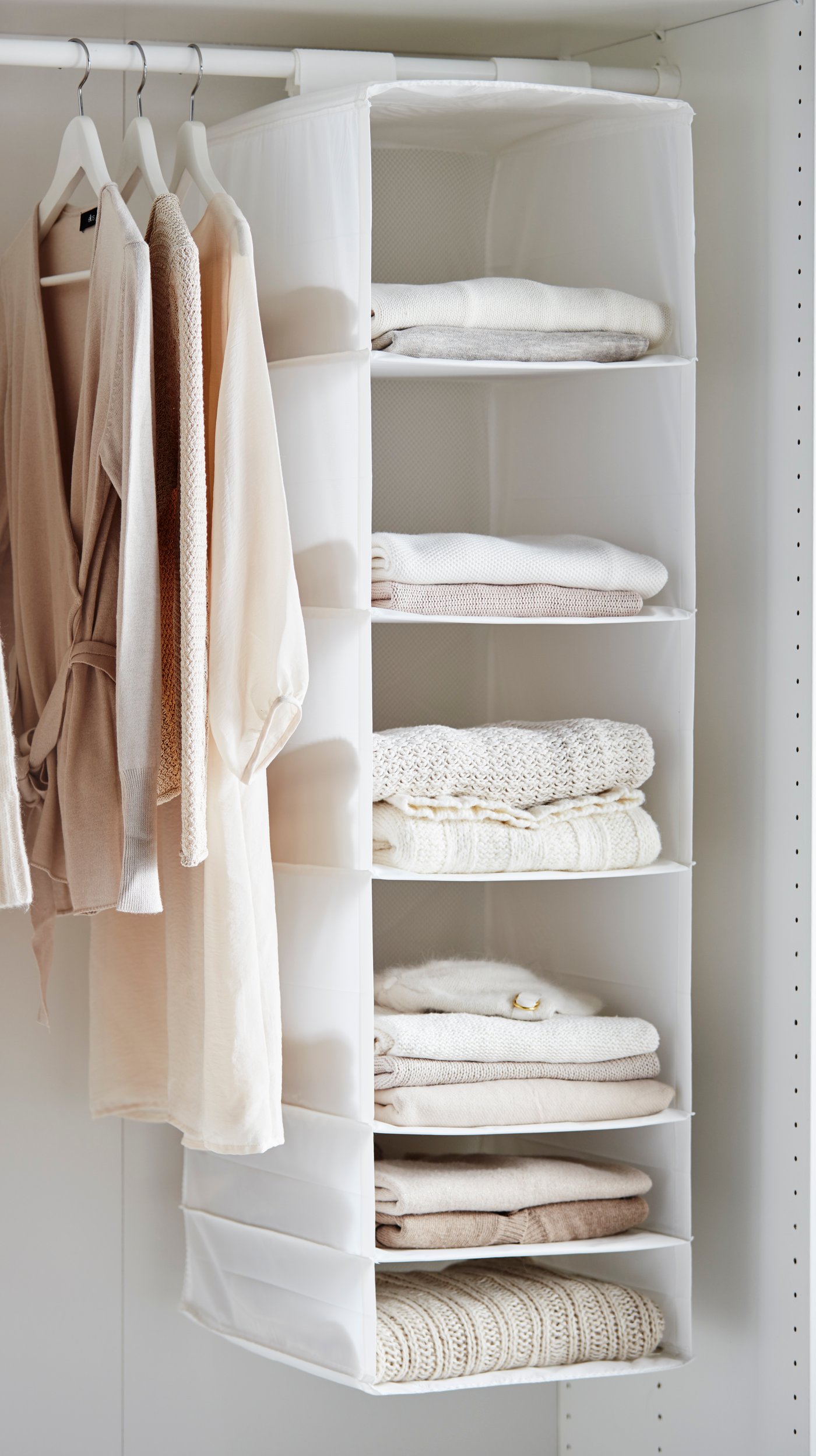 Open wardrobe with light clothing on hangers and a fabric shelf organizer holding neatly folded sweaters and towels.