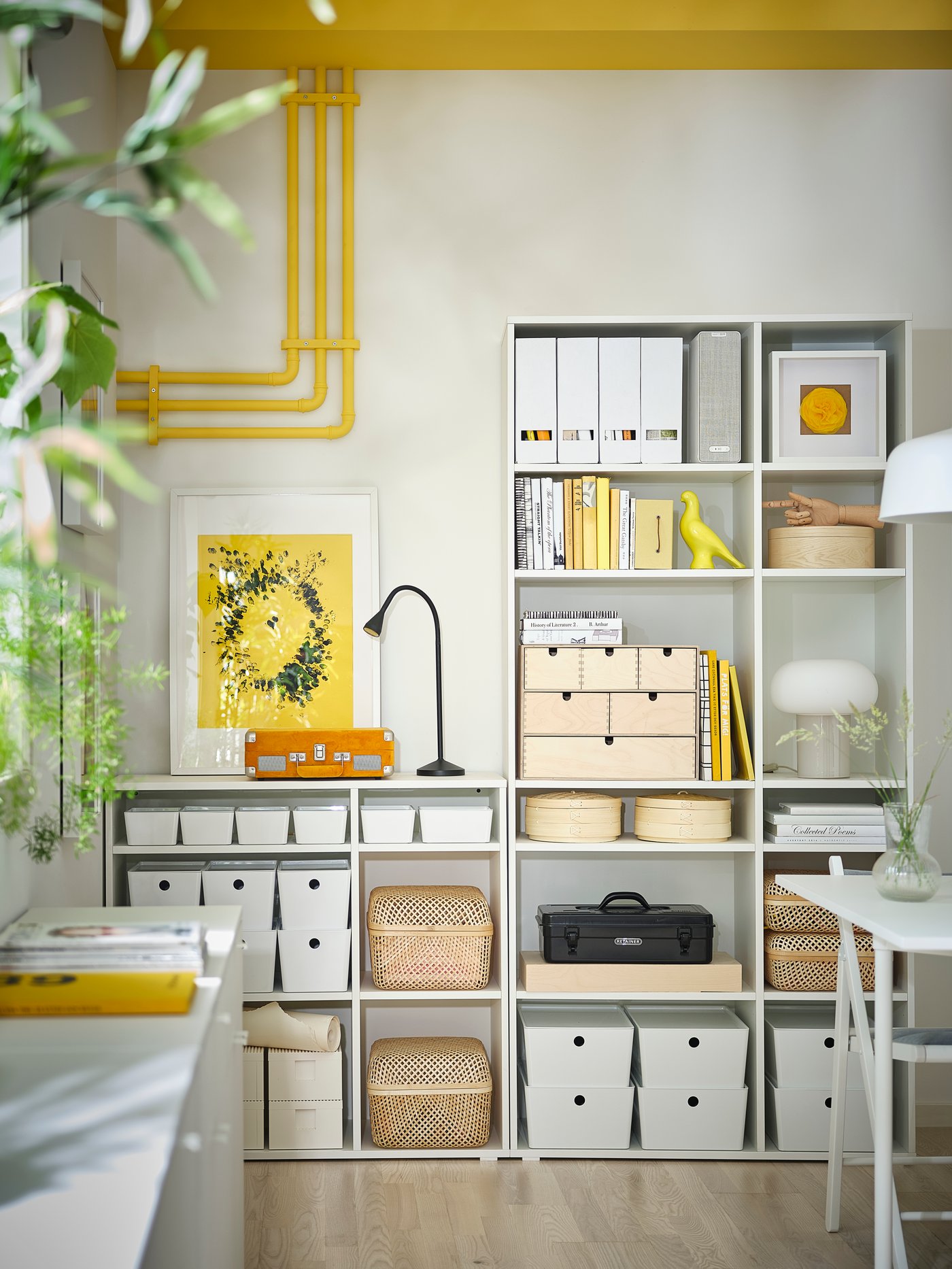 Organizovaný úložný prostor s policovým systémem plným bílých a béžových boxů, proutěných košů, knih a dekorací. Na stěně visí žlutý obraz, stropem vedou žluté trubky a na polici je černá stolní lampa. Interiér laděný do bílé, žluté a přírodního dřeva působí funkčně a esteticky.