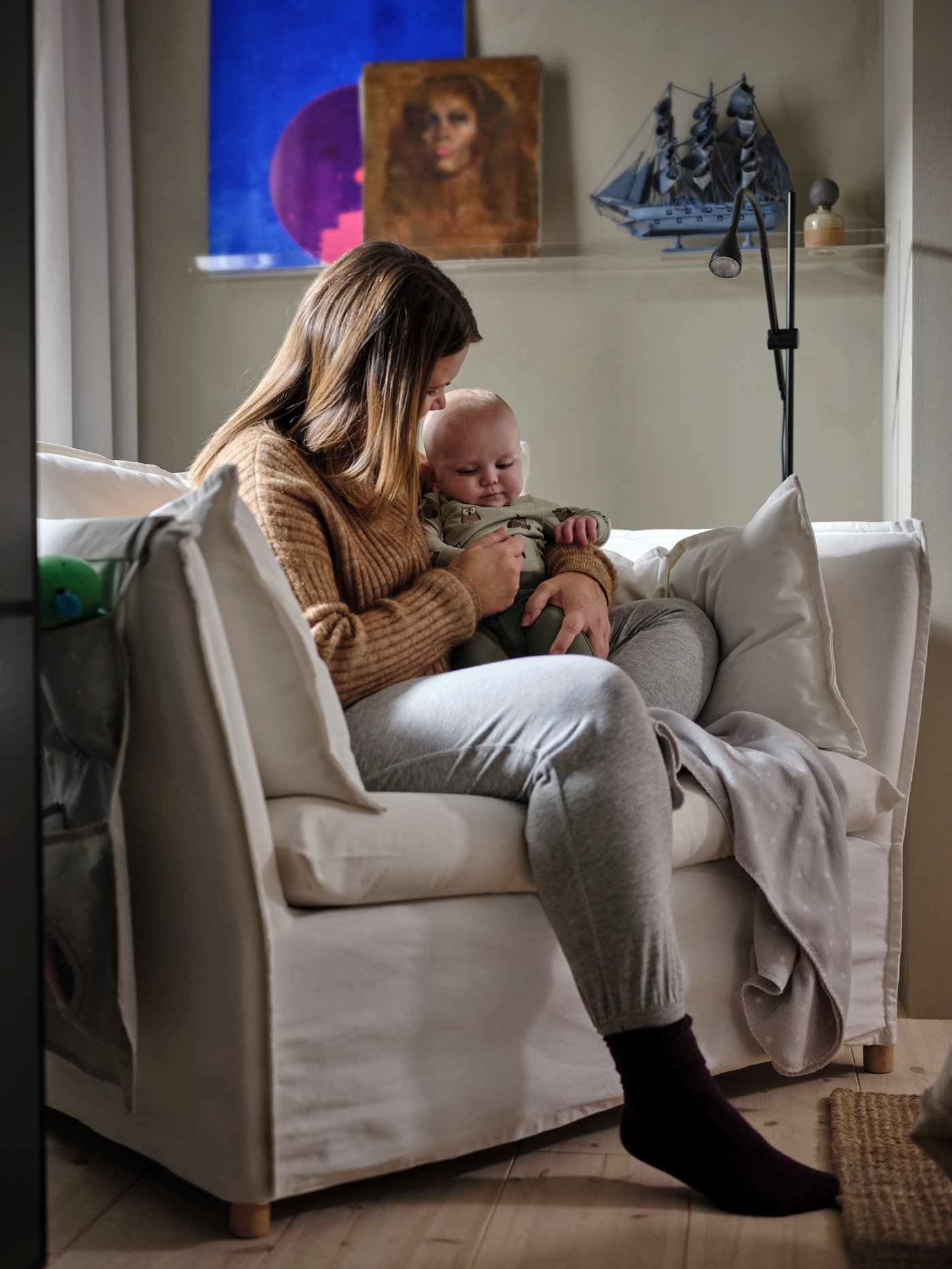 A woman sits on a white BACKSÄLEN 1.5 seater armchair holding a baby in her arms, with a LEN blanket on the seat beside her.