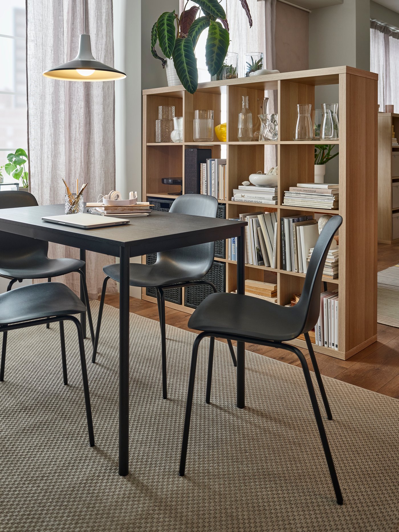 A black SANDSBERG dining table and LIDÅS chairs sit on a rattan rug, KALLAX shelving in oak effect acts as a room divider.