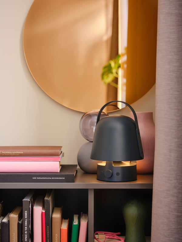 Plava VAPPEBY Bluetooth zvučnik-lampa na tamnosivom ormariću, pored knjiga, a ispred LED lampe za zid/ogledalo.