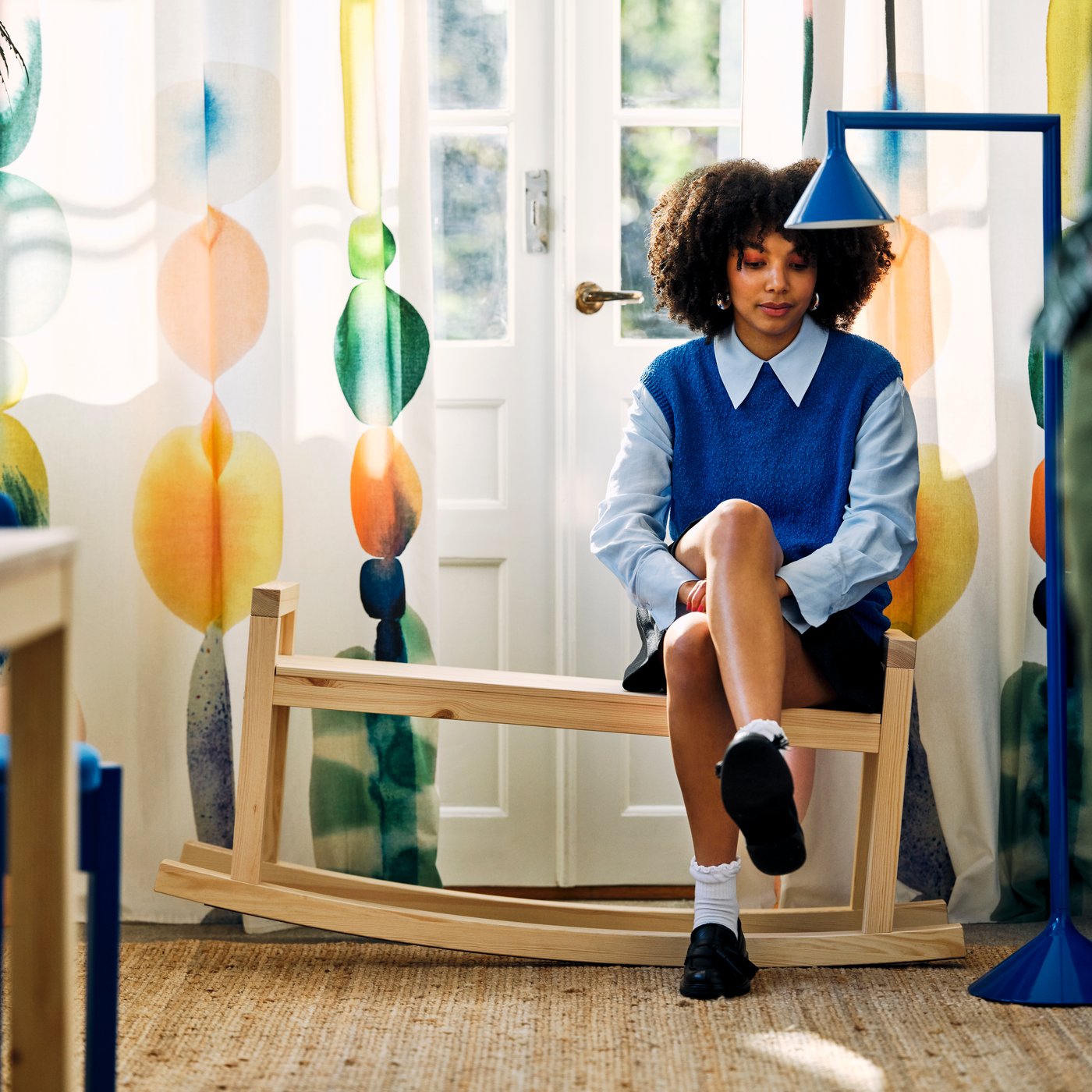 Person on wooden bench adjusts shoe near colorful curtains and white door.