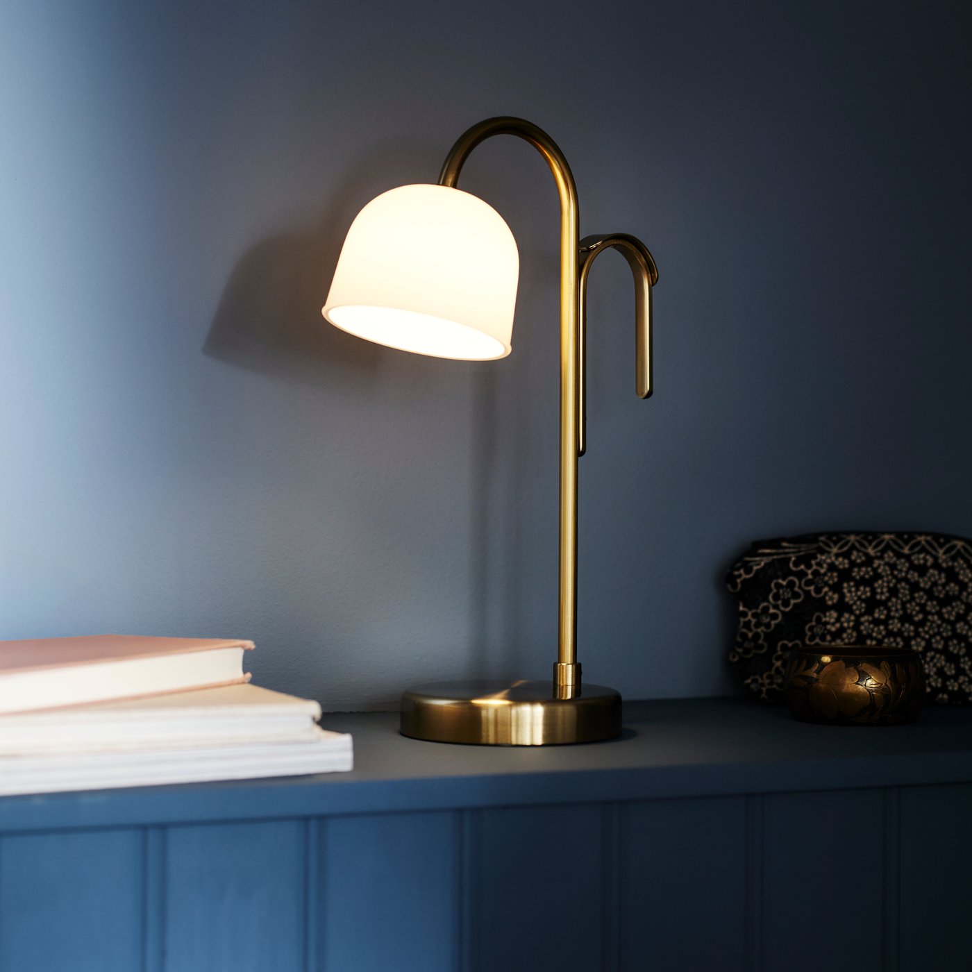 A brass/white glass TVÅMASTAD LED portable lamp that is switched on placed on a dark blue ledge with some books beside it.