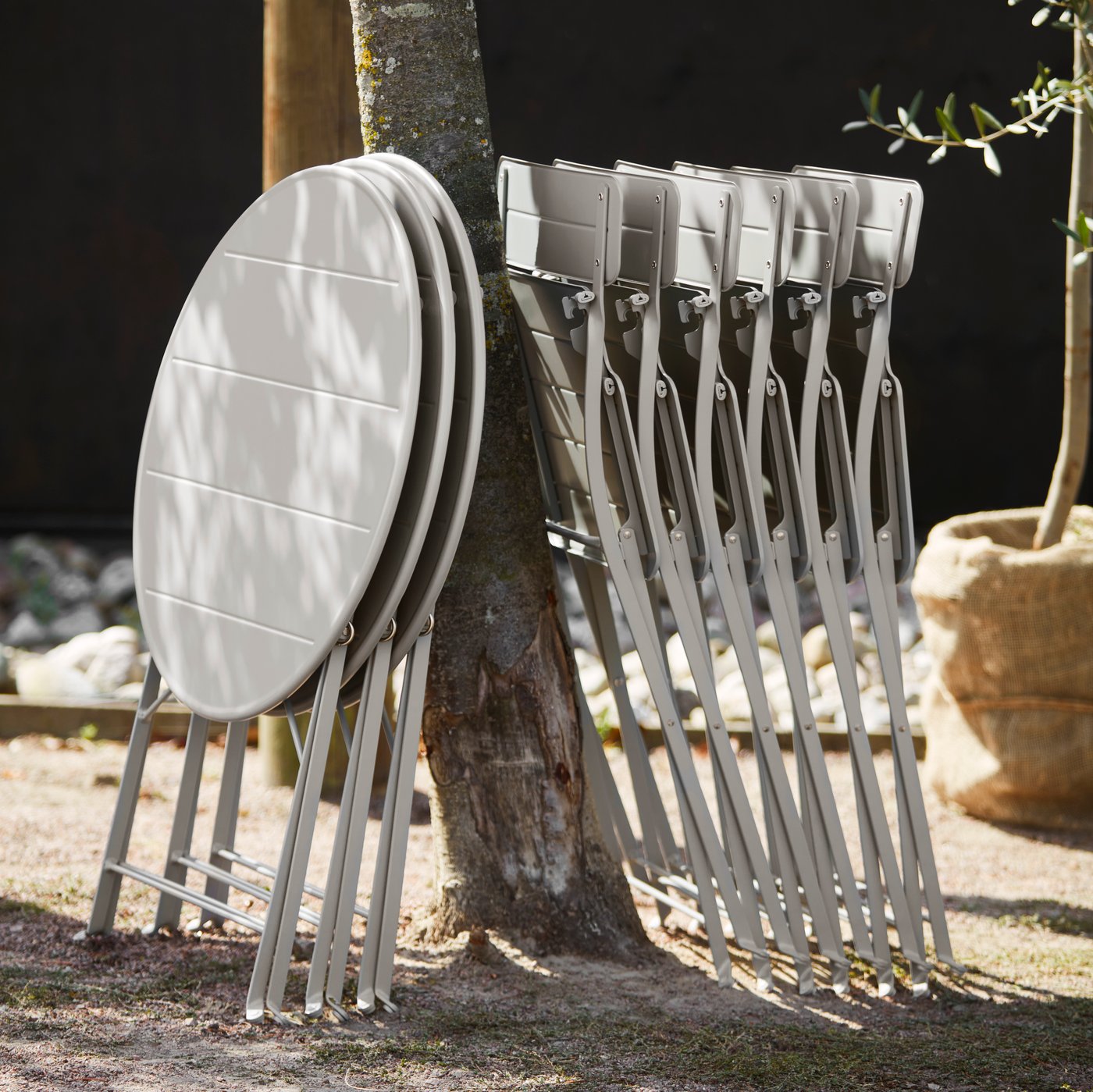 Plusieurs chaises et tables d’extérieur SUNDSÖ en blanc cassé sont appuyées de façon ordonnée contre un arbre.