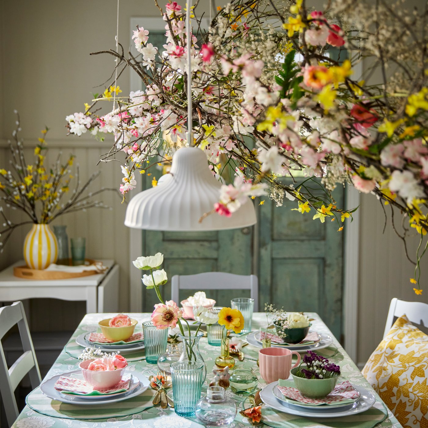 Some colorful SMÖRFISK mugs and UPPLAGA bowls sit on a beautifully set dining table adorned with flowers and greenery.