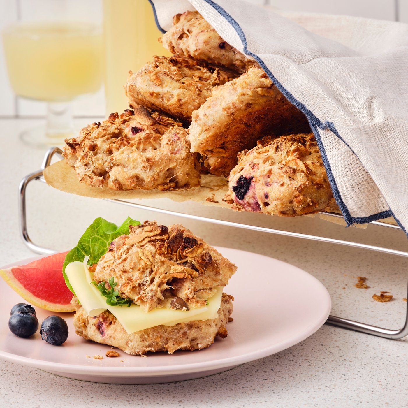 A granola breakfast bun with cheese and lettuce is placed on a pink plate and there are buns on a tray in the background.
